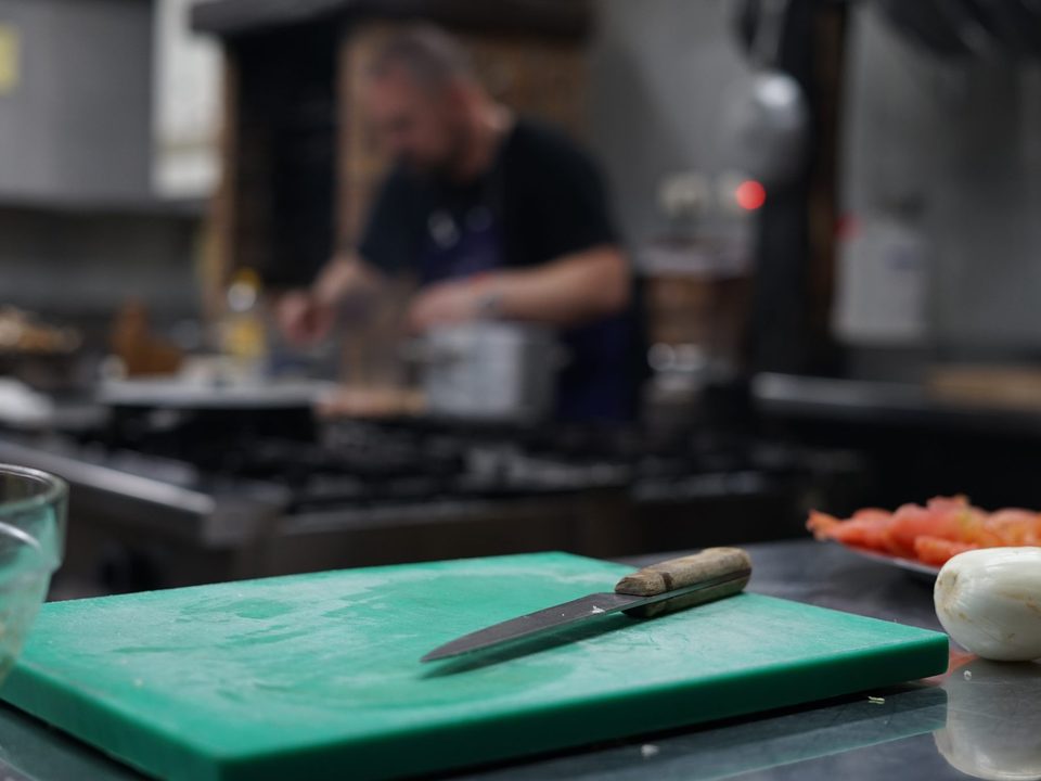 Chef-style kitchen prep with a knife on a green cutting board and a cook working at the stove in the background